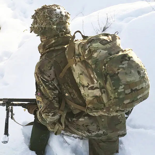 A person in full camouflage winter gear, including a helmet with netting, a backpack, and a jacket, crouching in deep snow while holding a rifle with a bipod.