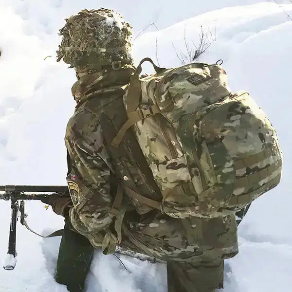 A person in full camouflage winter gear, including a helmet with netting, a backpack, and a jacket, crouching in deep snow while holding a rifle with a bipod.