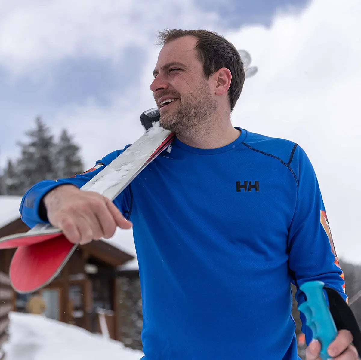 Man wearing blue HH Lifa Active Crew Long Sleeve Base Layer while carrying skis in snowy setting, showcasing winter performance at John Bulls.