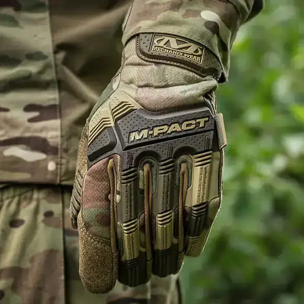 A close-up of a tactical glove worn on a hand, featuring a camouflage pattern with tan, brown, and olive green tones, and a prominent black and tan M-PACT Mechanical Wear protective panel on the back of the hand.