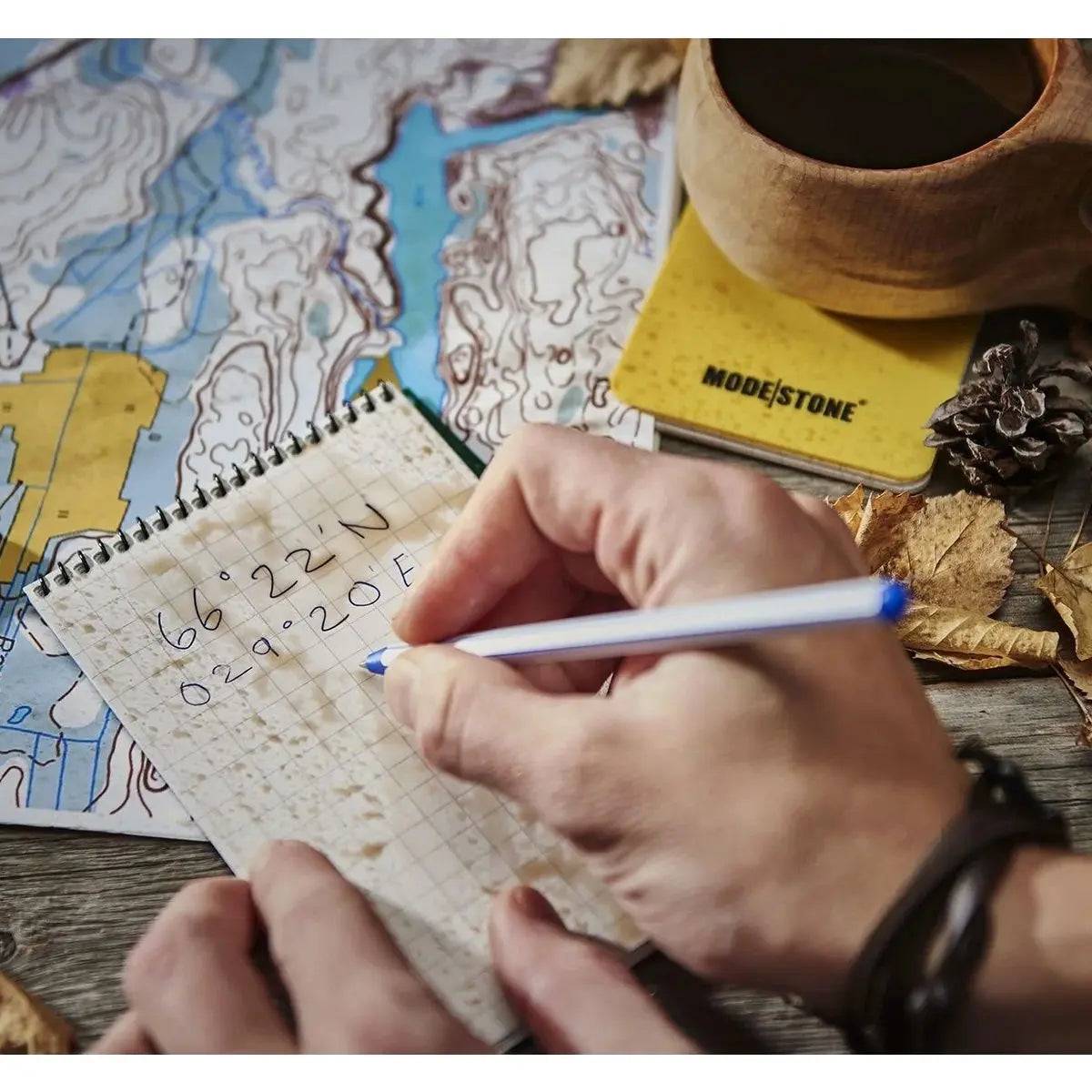 Person using the Modestone A6 Top Spiral Waterproof Notepad outdoors, writing coordinates on waterproof grid paper beside a map and coffee cup.