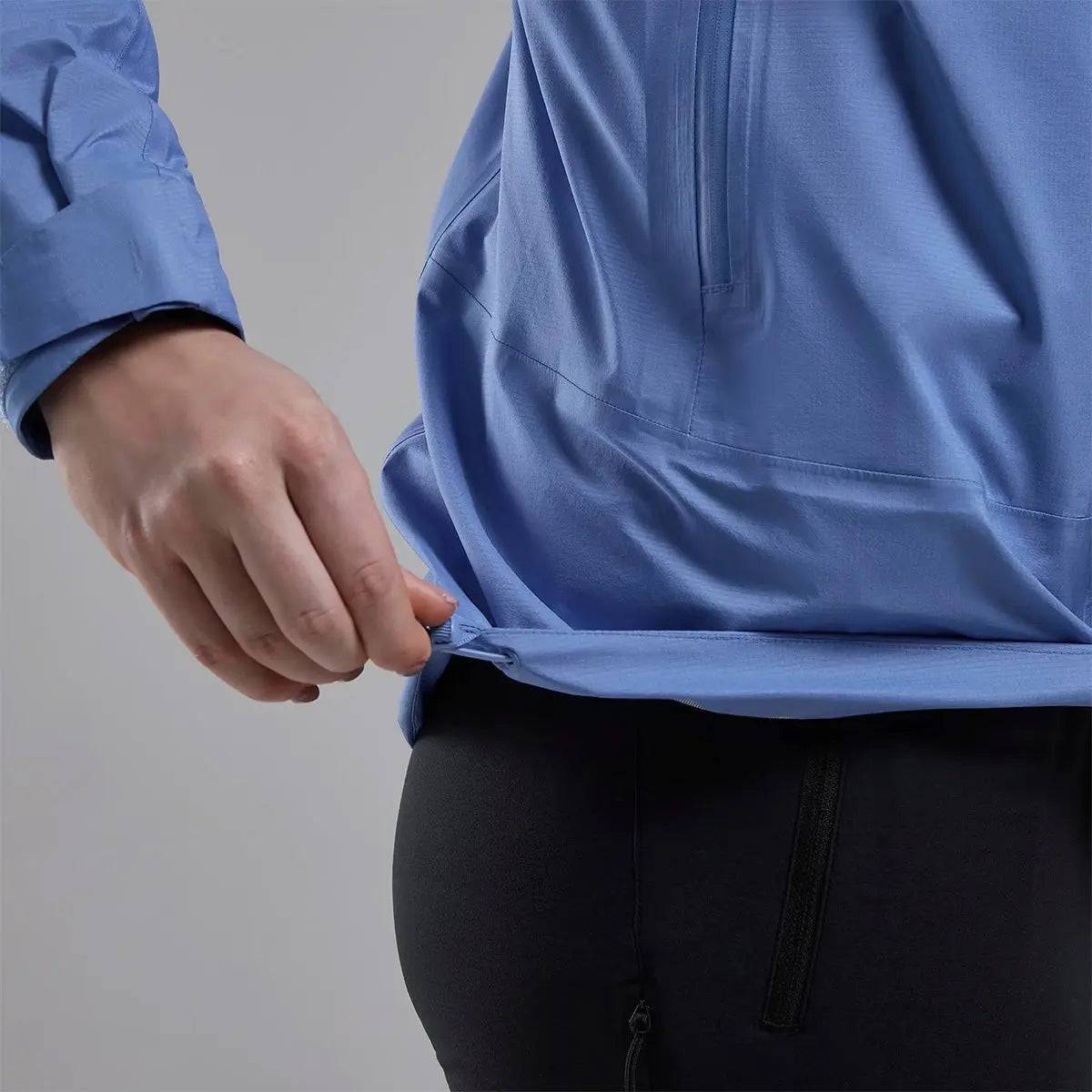 Model adjusting the hem of Montane Women's Cetus Lite Waterproof Jacket in light blue, demonstrating its customizable fit.