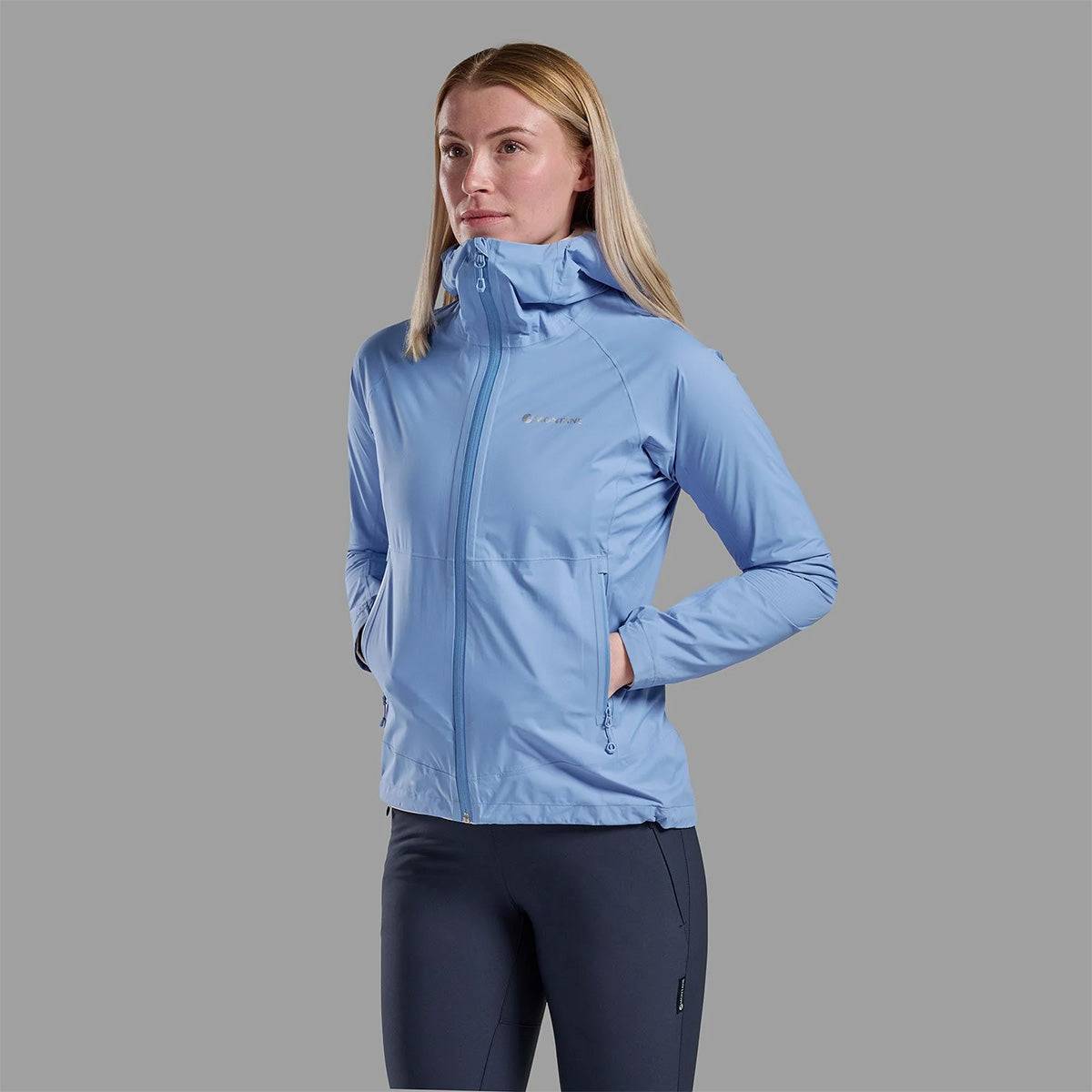 Model wearing sky blue Montane Womans Minimus Lite Waterproof Jacket with hands in pockets, showcasing stylish and functional rain coat for women.