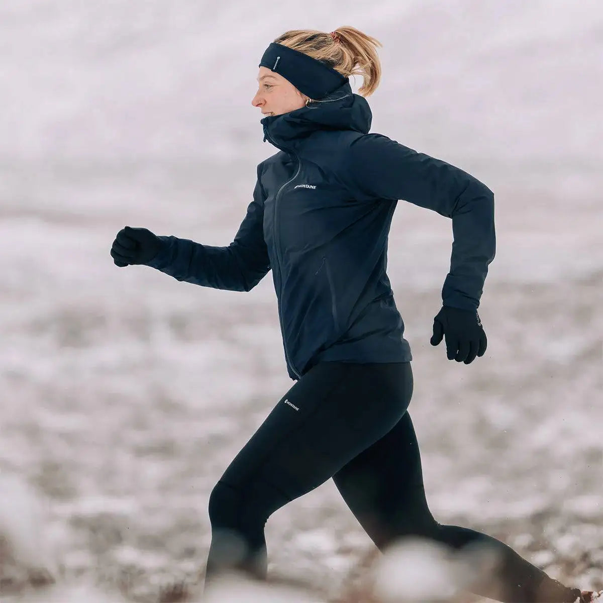 Woman trail running in snow wearing navy Montane Womans Minimus Lite Waterproof Jacket, built for high-performance in extreme weather conditions. from John Bull Clothing