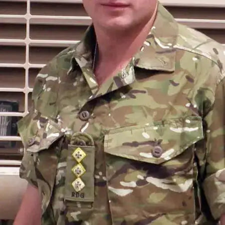 A close-up of a Captain of the RDG wearing a short-sleeve military camouflage uniform with a collar, chest pockets, and rank insignia on the left chest. The camouflage pattern mixes green, brown, tan, and black, and the background shows a beige slatted wall.