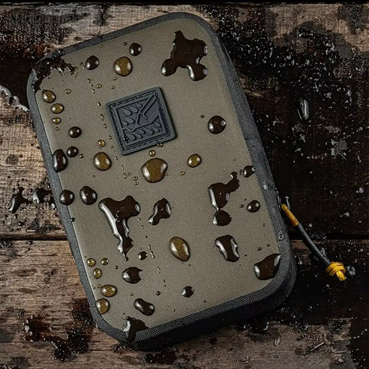 Rite in the Rain Monsoon Waterproof A6 Rain Shadow Pouch with water droplets on khaki fabric, black zipper, and yellow cord, on wooden surface