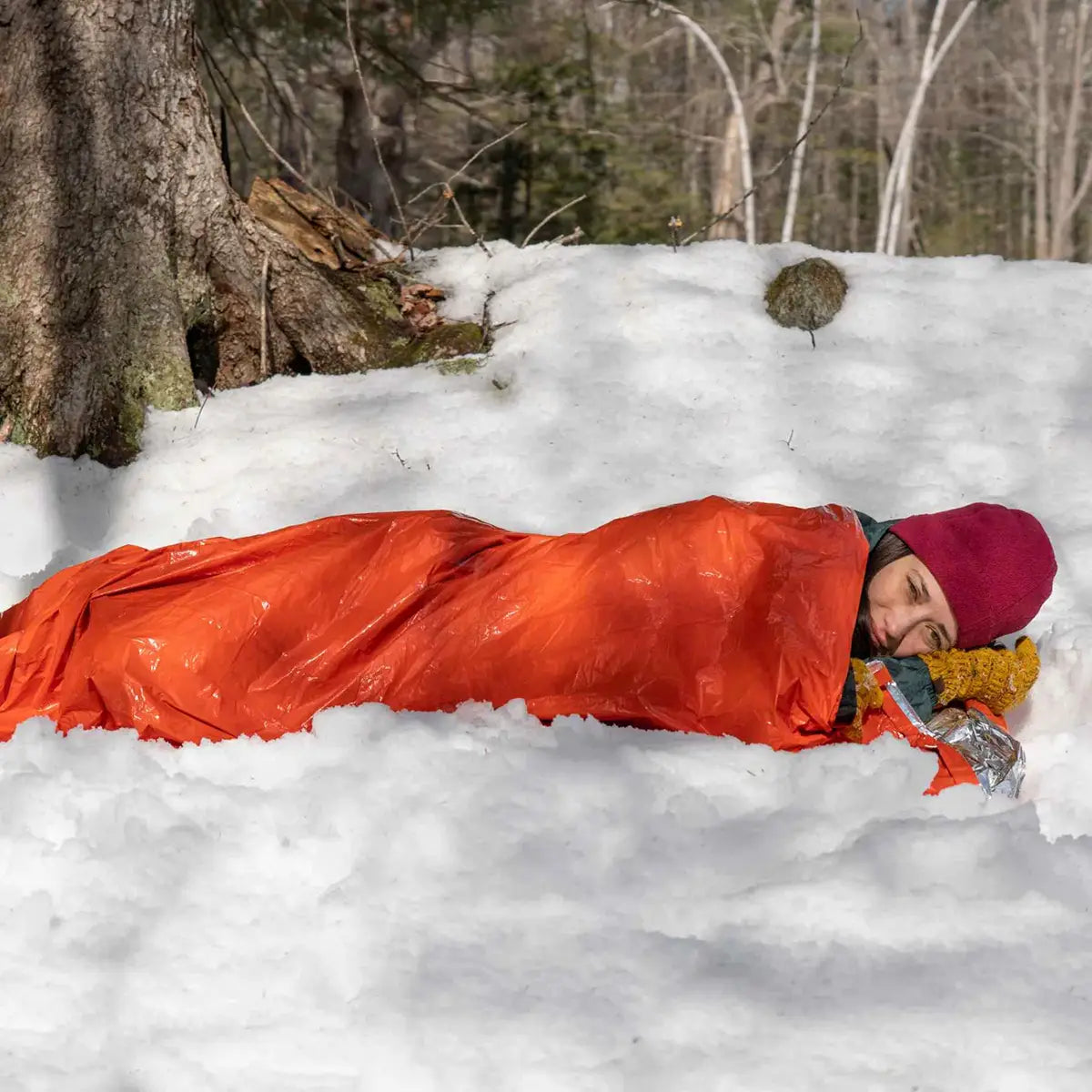 Camper sleeping in SOL Emergency Bivvy Bag Shelter with Whistle on snow, demonstrating insulation and emergency use.