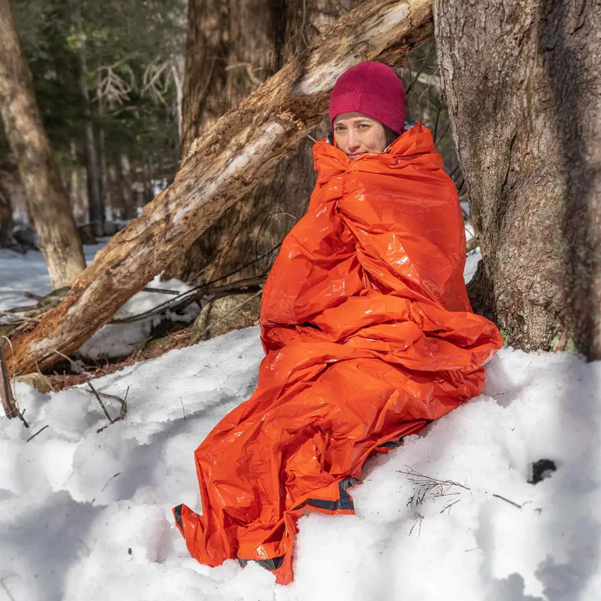 Person wrapped in SOL Emergency Bivvy Bag Shelter with Whistle, sitting in snow for cold-weather survival protection.