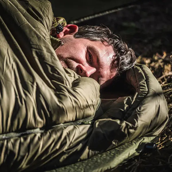 A man lying down wrapped in an olive green Snugpak sleeping bag or sleeping pad outdoors in dappled sunlight.