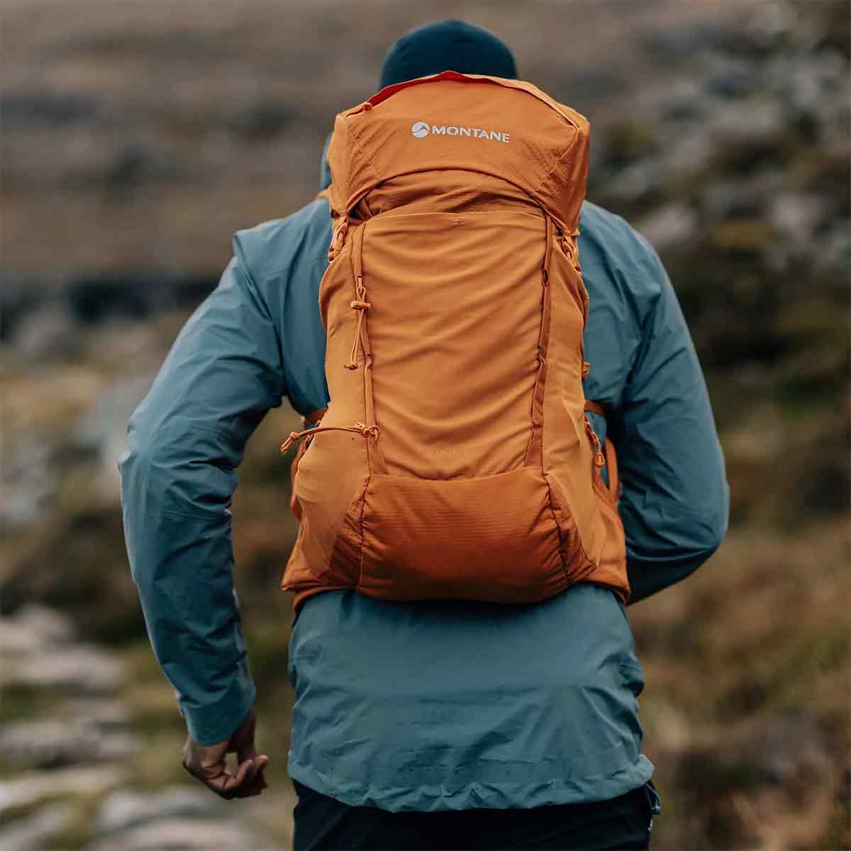 Hiker wearing the Montane Trailblazer 25L Rucksack in orange, trekking through rugged outdoor terrain with a secure fit.
