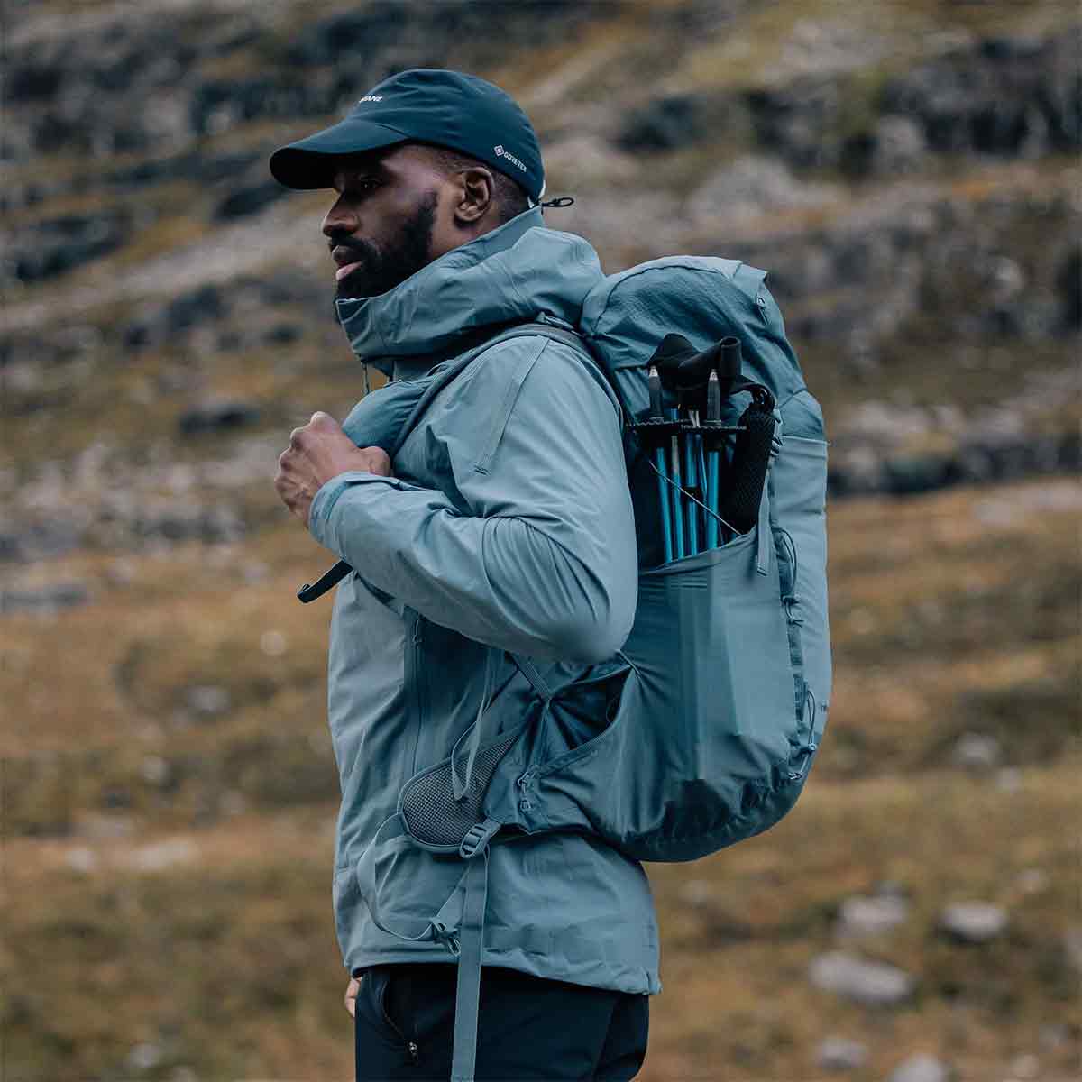 Hiker wearing the Montane Trailblazer 32 Litre Backpack in blue, demonstrating its ergonomic fit and side storage for trekking poles in a mountain setting.