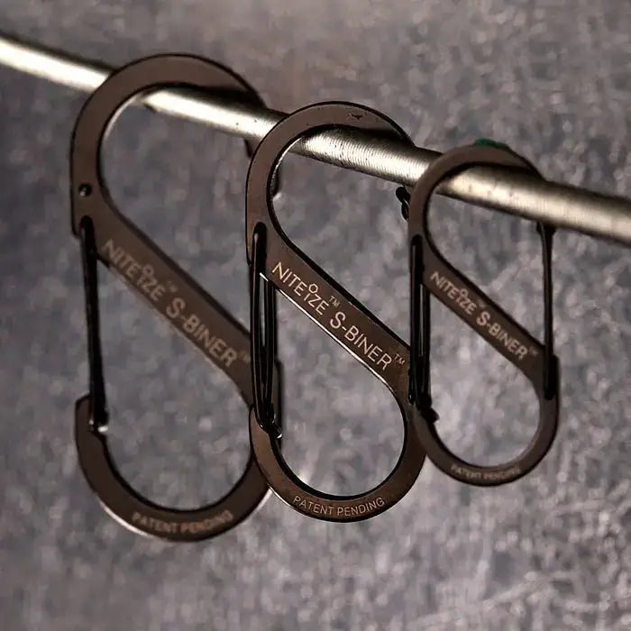 Nite-Ize S-Binner Size 3 Black Carabiner with dual spring gates hanging on a metal rod, ideal for camping and hiking gear organization. from John Bull Clothing