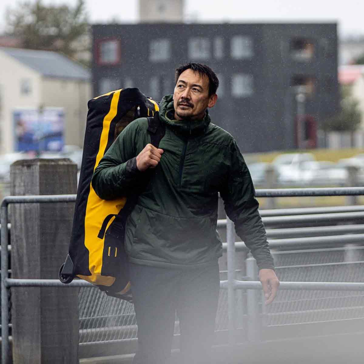 Man carrying Ortlieb Duffle RS Travel Bag 140L in black and yellow, demonstrating its portability in an outdoor setting.