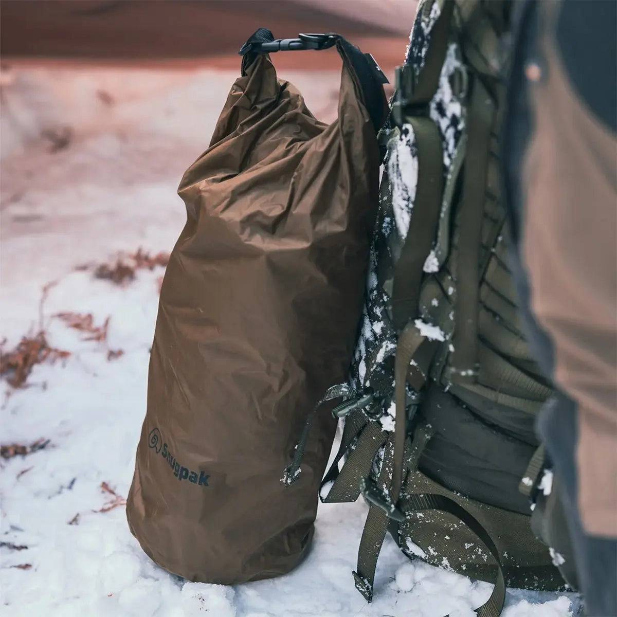Snugpak Dri-Sak Waterproof Dry Bag in brown, attached to a backpack in a snowy outdoor setting, ensuring dry storage in harsh conditions.