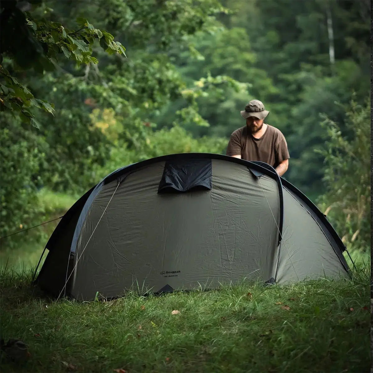 Camper setting up the Snugpak The Bunker IX Expedition Tent in a lush green outdoor environment, highlighting its ease of assembly.