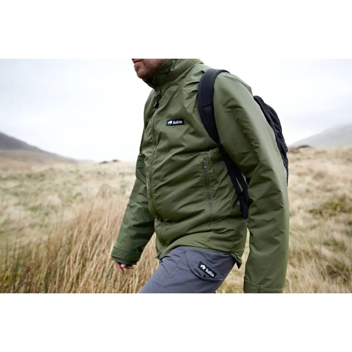 Man hiking in the Buffalo Mens Pertex Belay Jacket in olive green, showcasing its functionality in rugged outdoor terrain.