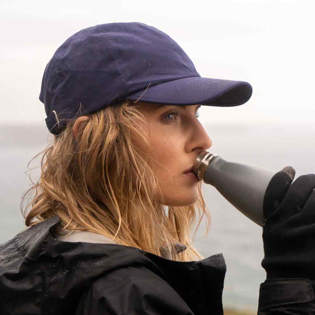 Woman wearing a navy Sealskinz Waterproof All Weather Baseball Cap while drinking from a bottle. Ideal for outdoor adventures in wet conditions.