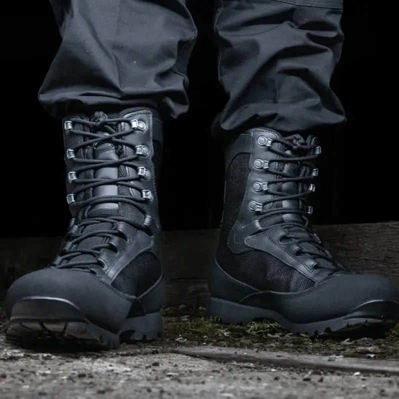 A close-up of a person wearing black AKU Pilgrim tactical boots with black pants, standing on a rough concrete surface against a dark background.