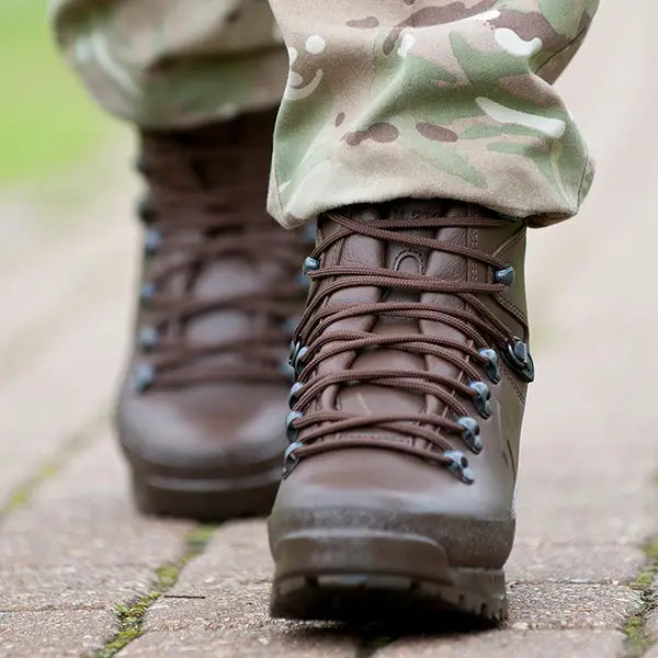 A close-up shot of a British Army Soldier wearing dark brown tactical boots with matching laces and camouflage pants, standing on a paved outdoor path.