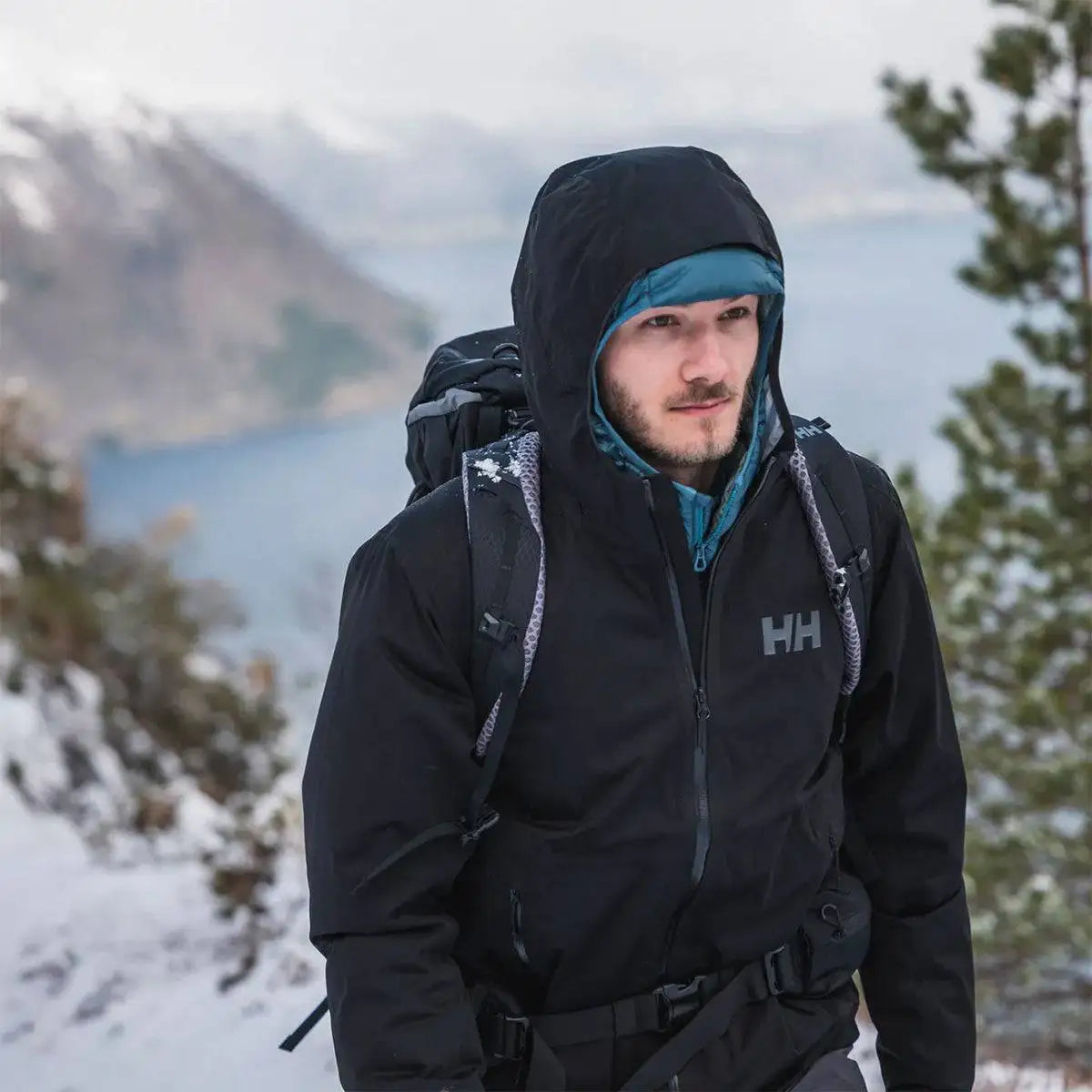 Man hiking in snowy mountains wearing black Helly Hansen Mens Momentum 3 Layer Stretch Waterproof Jacket, ideal for cold and wet conditions.  from  John Bull Clothing 