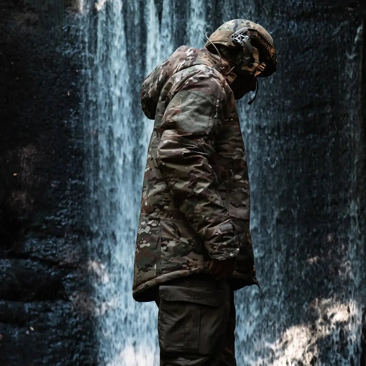 Person wearing Stoirm Mission Primaloft Waterproof Jacket Multicam in outdoor setting near waterfall, demonstrating rugged tactical use.