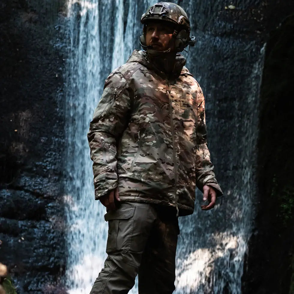 Model in Stoirm Mission Primaloft Waterproof Jacket Multicam standing in front of waterfall, showcasing field-ready design and camouflage.