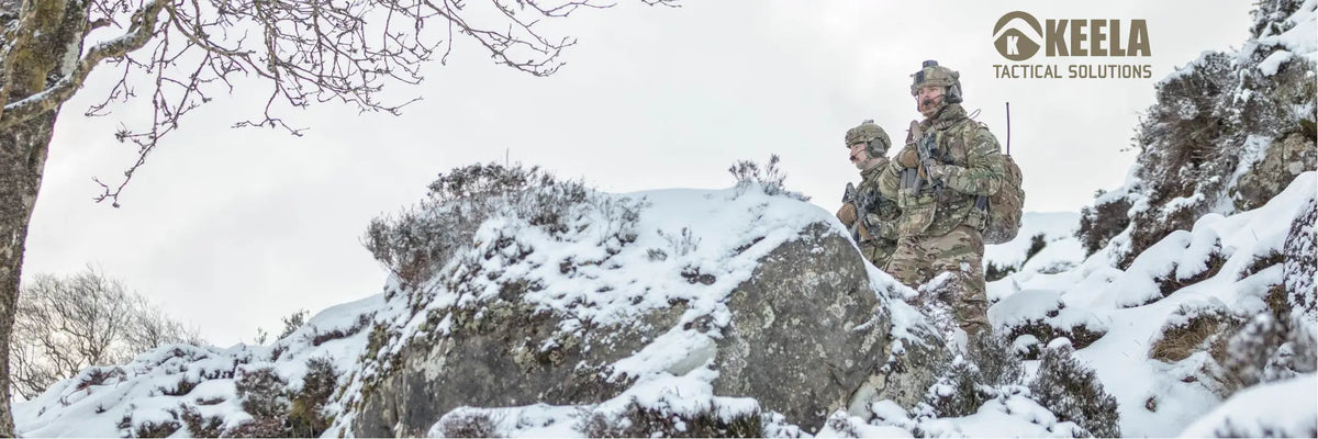 Two soldiers in camouflage winter gear standing on a snow-covered rocky hillside outdoors, with a cloudy sky and bare tree branches visible. The Keela Tactical Solutions logo appears in the top right corner.