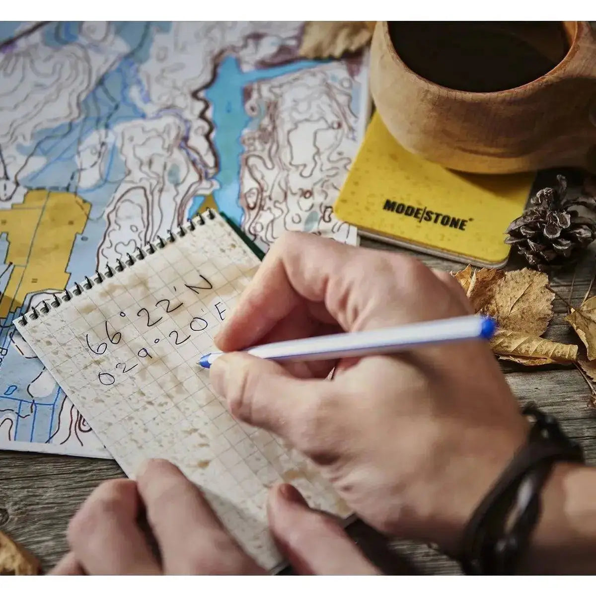 Person using the Modestone A6 Top Spiral Waterproof Notepad outdoors, writing coordinates on waterproof grid paper beside a map and coffee cup.  from  John Bull Clothing 