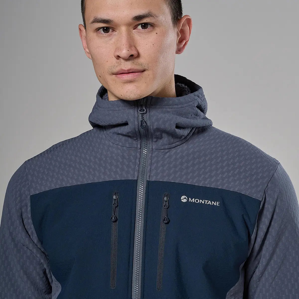 Male model wearing Montane Mens Dura XT Hoodie Fleece in navy blue and gray, featuring a high collar, front zipper, and multiple zippered pockets for outdoor activity.