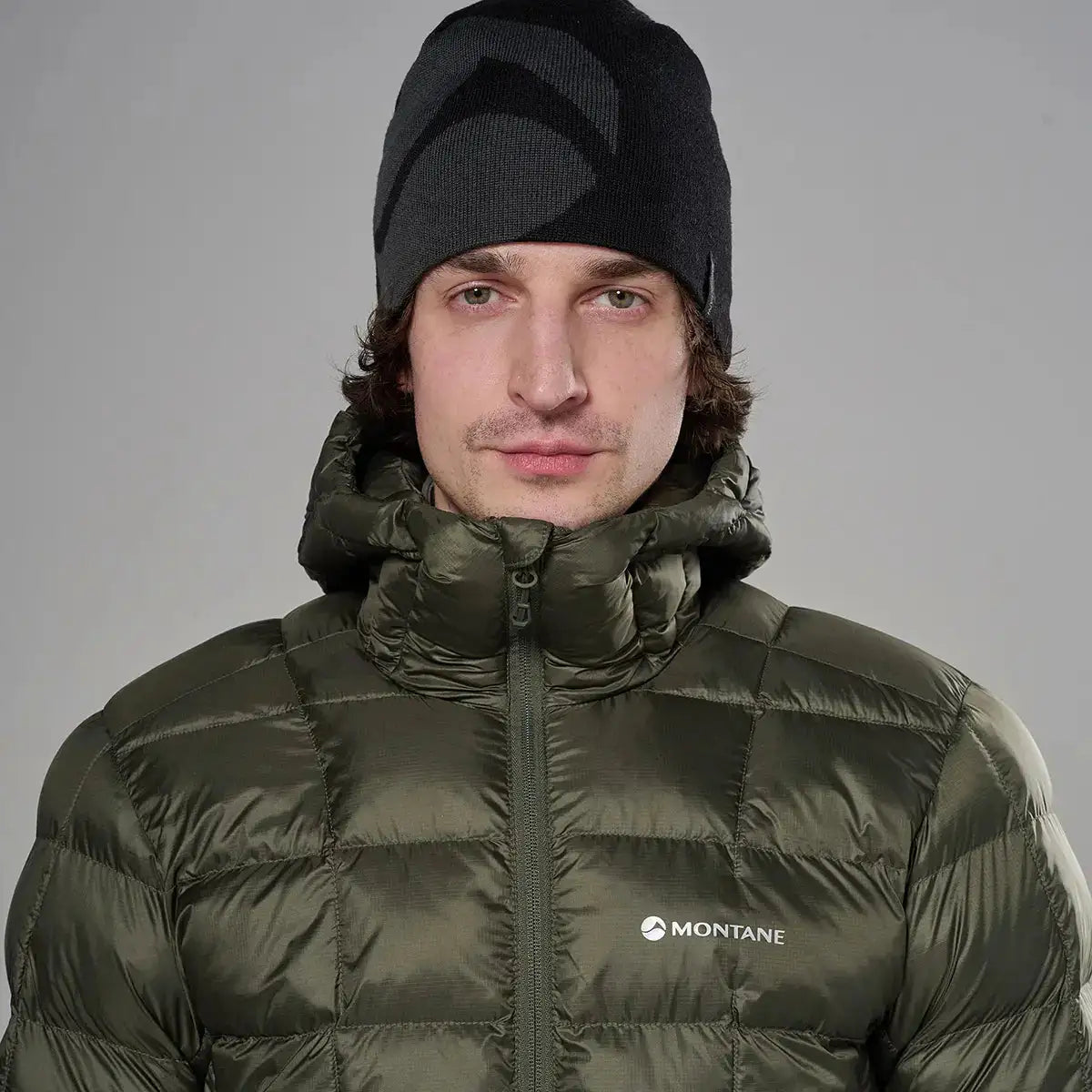 Male model wearing olive green Montane insulated puffer jacket with high collar and front zipper, paired with black beanie, on gray background