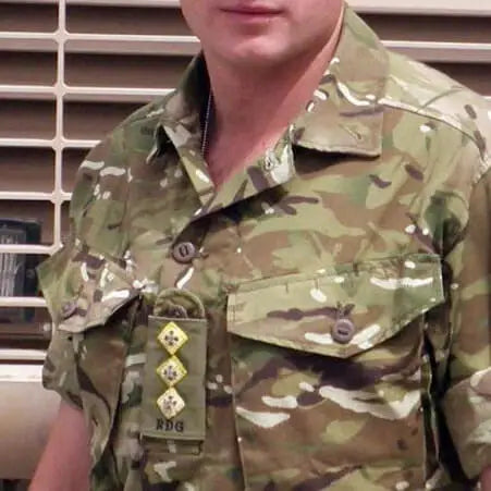 A close-up of a Captain of the RDG wearing a short-sleeve military camouflage uniform with a collar, chest pockets, and rank insignia on the left chest. The camouflage pattern mixes green, brown, tan, and black, and the background shows a beige slatted wall.