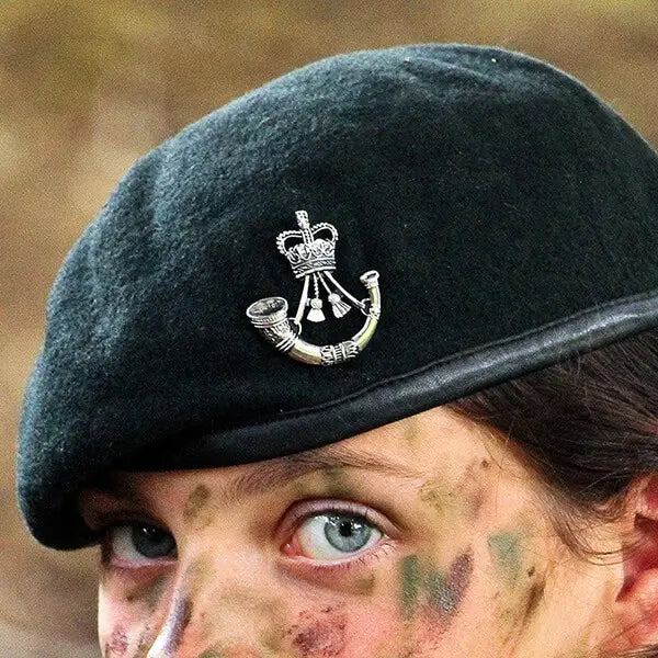 Close-up of a person wearing a Rifles Dark Green wool military beret with a silver Rifles Regiment cap badge with Queens Crown, featuring camouflage face paint.