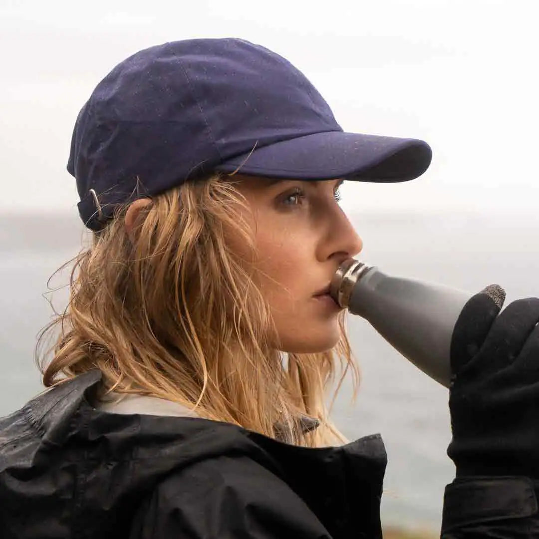 Woman wearing a navy Sealskinz Waterproof All Weather Baseball Cap while drinking from a bottle. Ideal for outdoor adventures in wet conditions.  from  John Bull Clothing 