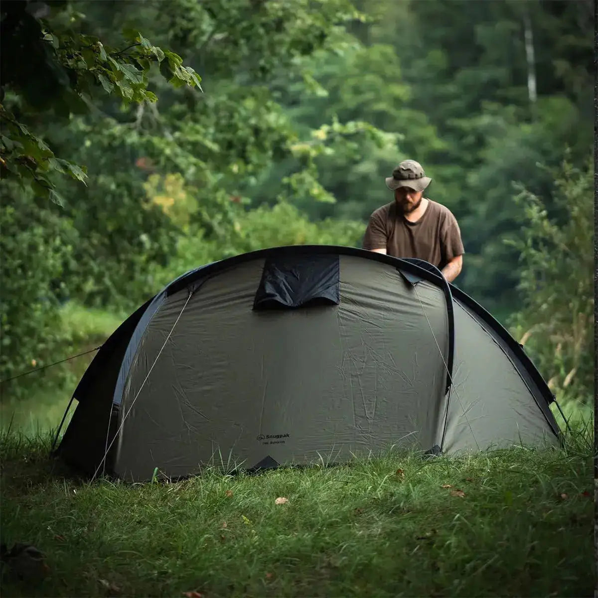 Camper setting up the Snugpak The Bunker IX Expedition Tent in a lush green outdoor environment, highlighting its ease of assembly.  from  John Bull Clothing 